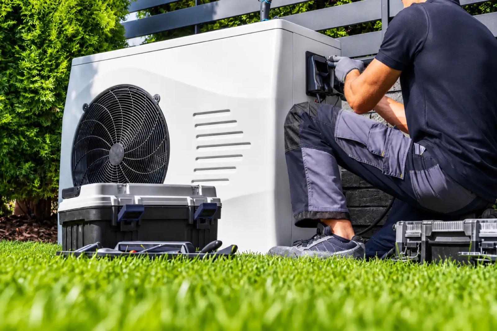 A technician kneeling beside a heat pump outdoor unit with a tool case on the grass