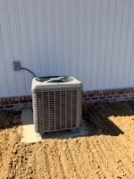 An outdoor air conditioning condenser on a small concrete pad beside a white metal building on bare dirt