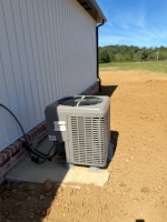 A new outdoor condenser on a pad beside a metal building with a green field and blue sky behind