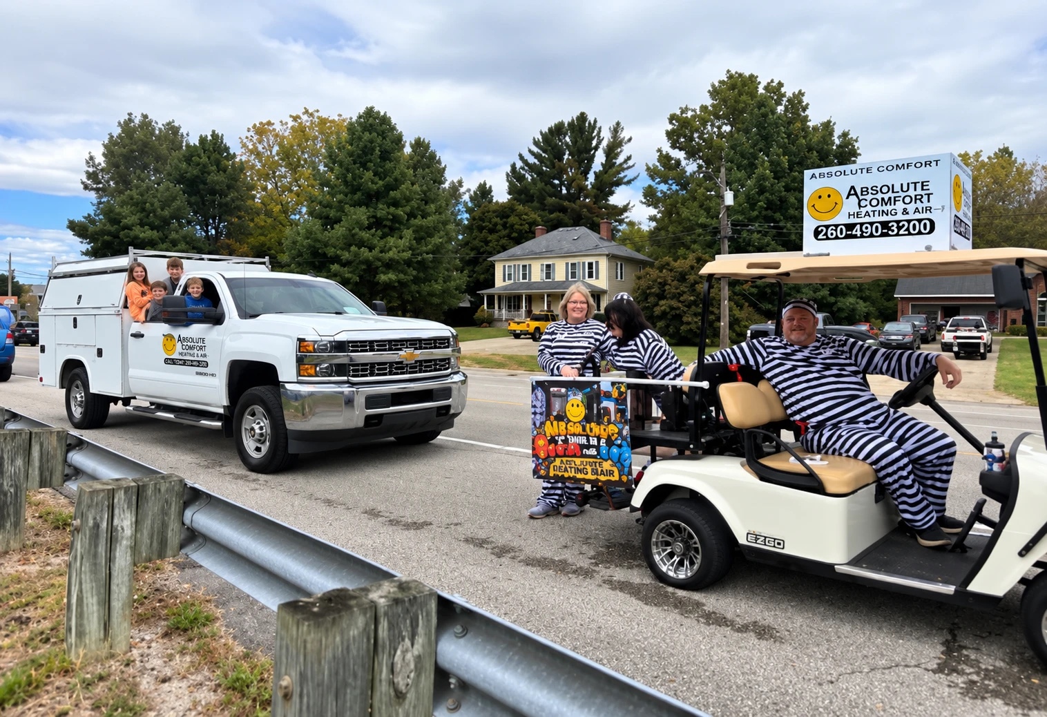 The white Absolute Comfort Heating & Air work truck with children in the back and a golf cart of people in striped costumes, with an Absolute Comfort sign, at a community event